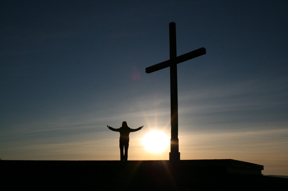 Silhouette of person with outstretched arms beside a large cross at sunset.